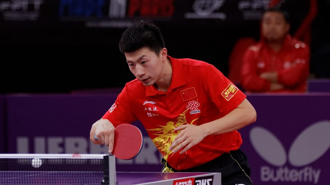 Ma Long in a red shirt playing a shot over the table against Koki Niwa