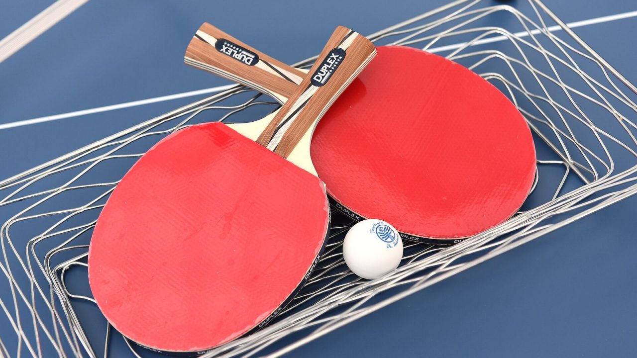 Two pre-made table tennis rackets with red rubbers and a white ball in a wire basket on a table
