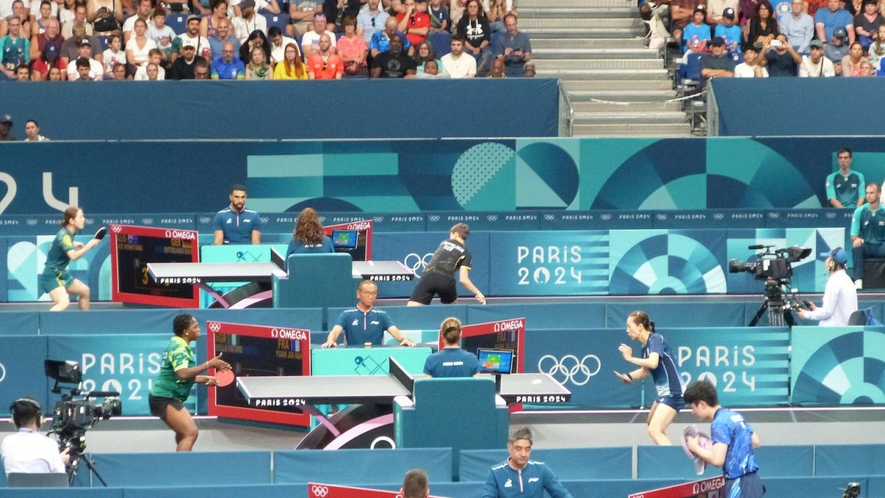 Table tennis players mid-rally on several tables at the South Paris Arena during the Paris 2024 Olympics