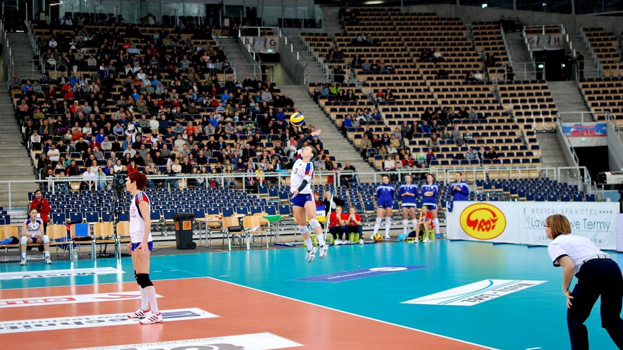 Courtney Thompson in mid-air hitting a jump serve from behind the end line in an indoor arena